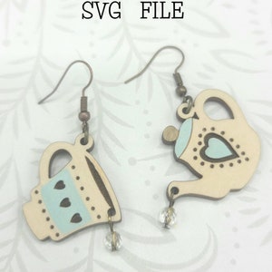 May include: A pair of wooden earrings featuring a teapot and a teacup design. The earrings are painted in light blue and white with a heart pattern. The earrings are labeled "Glowforge Ready SVG FILE" and "not a physical product".