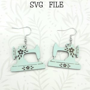 Puede incluir: Un par de pendientes de máquina de coser azul claro con detalles florales negros. Los pendientes están hechos de madera y están listos para ser cortados en una Glowforge. ARCHIVO SVG. no es un producto físico