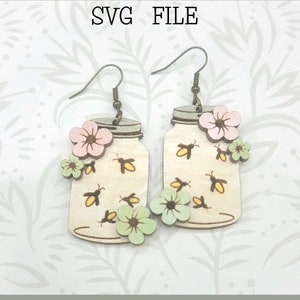 Può includere: File SVG pronto per Glowforge per orecchini. Gli orecchini sono in legno e presentano un barattolo di vetro con lucciole e fiori. I fiori sono rosa, verdi e gialli.