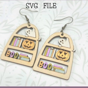 May include: A pair of wooden earrings shaped like bookshelves with a ghost and a jack-o-lantern on each shelf. The earrings are labeled "BOO" and have a gold and pink color scheme. Glowforge Ready SVG File. Not a physical product.