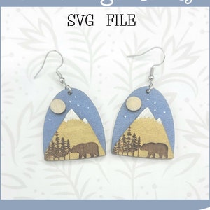 May include: A pair of wooden earrings with a blue and brown mountain scene, a white moon, and a bear silhouette. The earrings are labeled "Glowforge Ready SVG FILE" and "not a physical product".