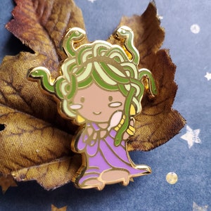 May include: A gold-coloured enamel pin depicting a cartoon Medusa with green snakes for hair, wearing a purple dress. The pin has a gold outline and is set against a brown leaf.