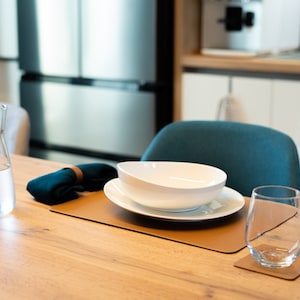 May include: A table setting with a white bowl, a plate, a glass, and a jug of water. The table is made of wood and has a brown leather placemat.