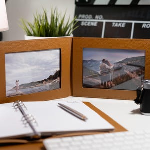 May include: Two brown leather photo frames with a white mat and glass front. The frames are hinged together and open to reveal two photos. One photo shows a couple standing on a cliff overlooking a body of water. The other photo shows two people standing on a beach.