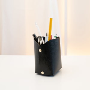 May include: A black leather pencil holder with a variety of pens and pencils inside. The holder is sitting on a white surface.