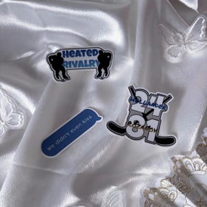 May include: A collection of stickers with various designs. One sticker reads "Heated Rivalry" with hockey player silhouettes. Another says "24 81 Hollander & Randy" with hockey sticks. A blue speech bubble says "we didn't even kiss".