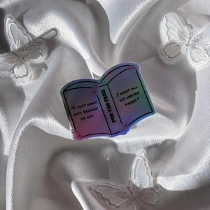May include: A sticker in the shape of an open book with the text "I JUST WANT HIM, BROKEN OR HOT" and "I WANT ALL HIS UNEVEN PIECES." The sticker has a holographic effect and is surrounded by white butterflies on a white satin background.