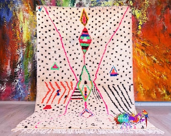 Pode incluir: Um tapete marroquino branco com um padrão geométrico em rosa, verde, azul e laranja. O tapete tem uma borda de franjas e é feito de lã.