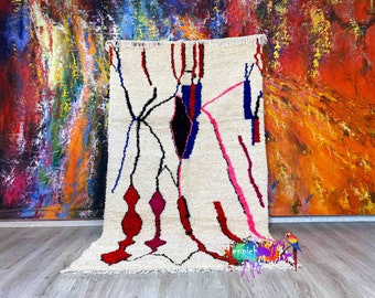 Peut inclure: Tapis crème avec un motif géométrique composé de lignes et de formes rouges, roses, noires et bleues. Le tapis a une surface texturée et une frange sur un bord. Le fond est une peinture abstraite colorée.