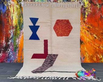 Op de afbeelding: Een wit tapijt met een geometrisch ontwerp met een rode zeshoek, een blauwe driehoek en een rode en bruine L-vormige figuur. Het tapijt heeft een franje rand.
