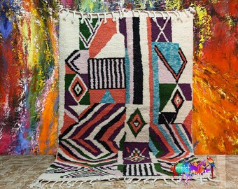 Peut inclure: Un tapis marocain coloré avec des motifs géométriques dans des tons de rose, bleu, vert et noir. Le tapis a un fond blanc et est tissé avec une bordure à franges.