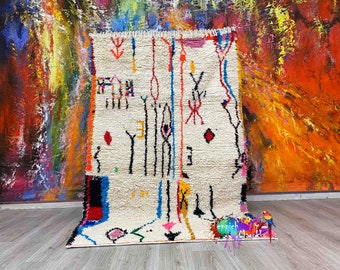 Peut inclure: Tapis rectangulaire crème avec un motif géométrique composé de lignes et de formes noires, rouges, bleues et jaunes. Le tapis a une frange sur le bord supérieur et est présenté sur un sol en bois clair. Le fond est une peinture abstraite colorée.