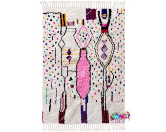 Peut inclure: Tapis rectangulaire blanc cassé avec un motif abstrait coloré. Le tapis présente un motif central rose en forme de bouteille, entouré de formes géométriques dans diverses couleurs, dont le rose, le jaune et le bleu. Bordure à franges.