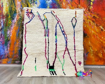Peut inclure: Un tapis marocain blanc avec un motif géométrique en noir, rose, bleu et vert. Le tapis a une bordure frangée et est fait de laine.