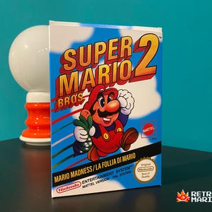 Può includere: Una scatola del videogioco Super Mario Bros. 2 con Mario che tiene in mano una verdura verde. La scatola ha uno sfondo di cielo blu con il titolo del gioco in giallo e rosso. Include anche il testo "MARIO MADNESS/LA FOLLIA DI MARIO".