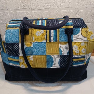 May include: A quilted tote bag with a patchwork design featuring blue, yellow and white fabric. The bag has two black handles.