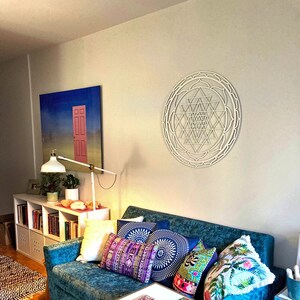 May include: A white wooden Sri Yantra mandala wall hanging with intricate geometric designs. The mandala is hanging on a white wall above a teal blue couch with colorful throw pillows.