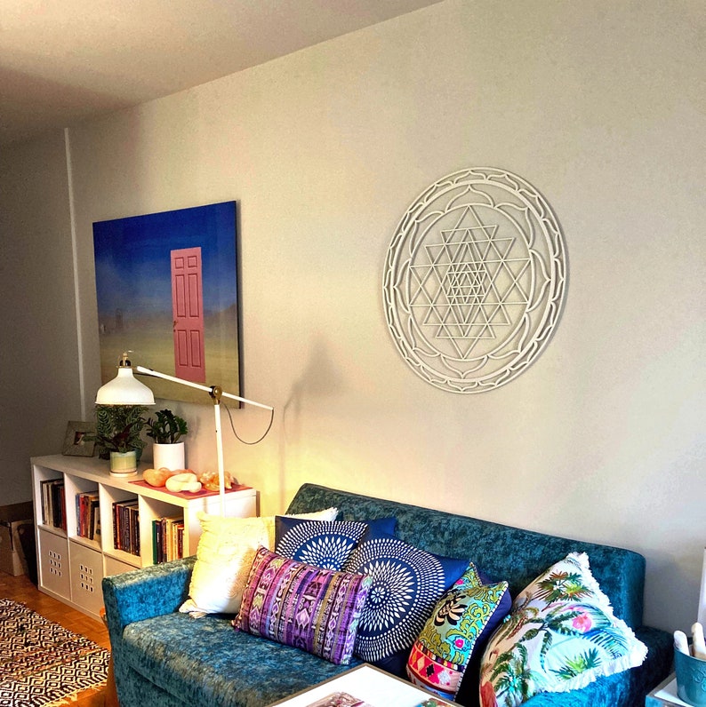 May include: A white wooden Sri Yantra mandala wall hanging with intricate geometric designs. The mandala is hanging on a white wall above a teal blue couch with colorful throw pillows.