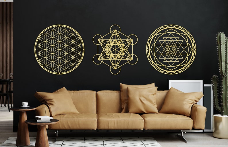 May include: Three gold geometric wall decor pieces, including a flower of life, a metatron's cube, and a sri yantra, are displayed on a black wall above a brown leather couch.