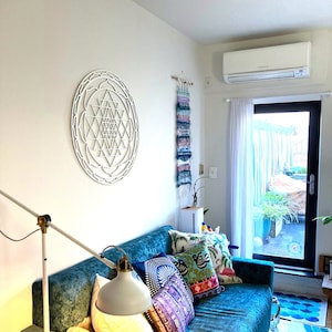 May include: A white wall with a large white geometric wall hanging. A blue couch with colorful pillows is in front of the wall. The couch is in a living room with a window and a white air conditioner.