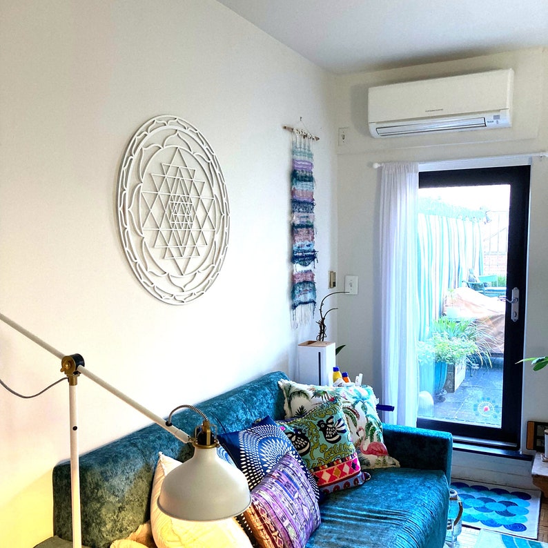 May include: A white wall with a large white geometric wall hanging. A blue couch with colorful pillows is in front of the wall. The couch is in a living room with a window and a white air conditioner.