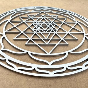 May include: A silver metal Sri Yantra mandala with a detailed geometric design. The mandala is circular and has a decorative border.