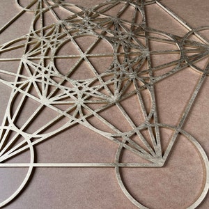 May include: A gold-colored, geometric wall art piece featuring intersecting circles, triangles, and lines. The artwork is set against a neutral, light brown background, creating a striking contrast. The design is intricate and symmetrical.