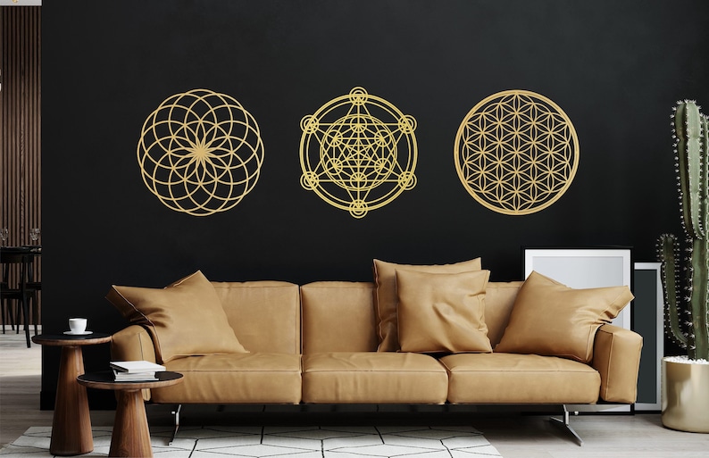May include: Three gold geometric wall decor pieces in the shape of a flower of life, a metatron's cube, and another flower of life. The decor is hanging on a black wall above a brown leather couch.