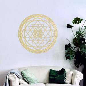 May include: A gold-colored geometric mandala wall decor with a circular design. The mandala is made of intricate triangles and squares, and it is surrounded by a decorative border. The mandala is hanging on a white wall in a living room setting.