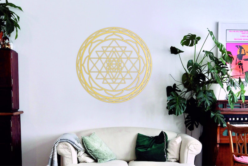 May include: A gold-colored geometric mandala wall decor with a circular design. The mandala is made of intricate triangles and squares, and it is surrounded by a decorative border. The mandala is hanging on a white wall in a living room setting.