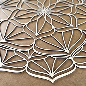 Op de afbeelding: Een witte houten mandala met een geometrisch ontwerp. De mandala is gemaakt van meerdere lagen hout die zijn uitgesneden om een ingewikkeld patroon te cre&euml;ren.