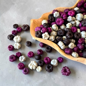Könnte beinhalten: Eine Sammlung kleiner, dekorativer kürbisförmiger Ornamente in den Farben Dunkelviolett, Schwarz und Cremeweiß. Viele dieser Miniaturkürbisse befinden sich in einer großen, hellen Holzschale, während andere in der unmittelbaren Umgebung verstreut sind.