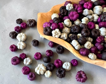 Puede incluir: Una colección de pequeños adornos decorativos con forma de calabaza en tonos morado intenso, negro y blanco roto. Muchas de estas calabazas en miniatura están contenidas dentro de un cuenco de madera de color claro, mientras otras están esparcidas.