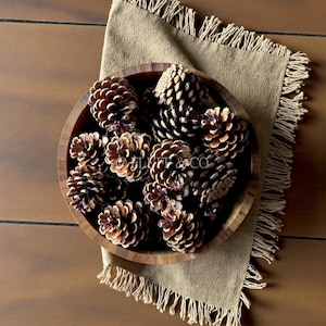 Puede incluir: Un cuenco de madera lleno de piñas marrones, sobre un paño beige con flecos. Las piñas tienen una superficie texturizada y diferentes tonos de marrón. El cuenco está sobre una superficie de madera.