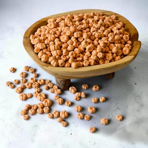 Puede incluir: Un cuenco grande y ovalado de madera marrón claro con tres patas cortas, lleno de una multitud de pequeños adornos de color naranja mate con forma de calabaza. Varias de estas figuras de calabaza en miniatura están esparcidas fuera.