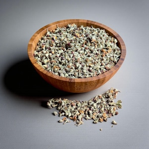 May include: A wooden bowl filled with dried herbs, with a small pile of herbs next to the bowl. The herbs are a mix of green, brown, and orange. The bowl is made of light brown wood and sits on a grey surface.