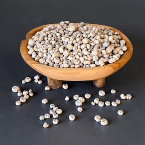 Puede incluir: Un cuenco redondo de madera marrón claro con tres patas cortas, lleno hasta el borde con una gran cantidad de pequeñas esferas botánicas secas, de color blanco roto y estriadas. Varios de estos objetos secos y texturizados están esparcidos.