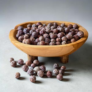May include: A round, light-brown wooden bowl with three short, sturdy legs, filled with a large quantity of dried, dark purple-brown juniper berries. Several additional dried juniper berries are scattered around the base of the bowl.