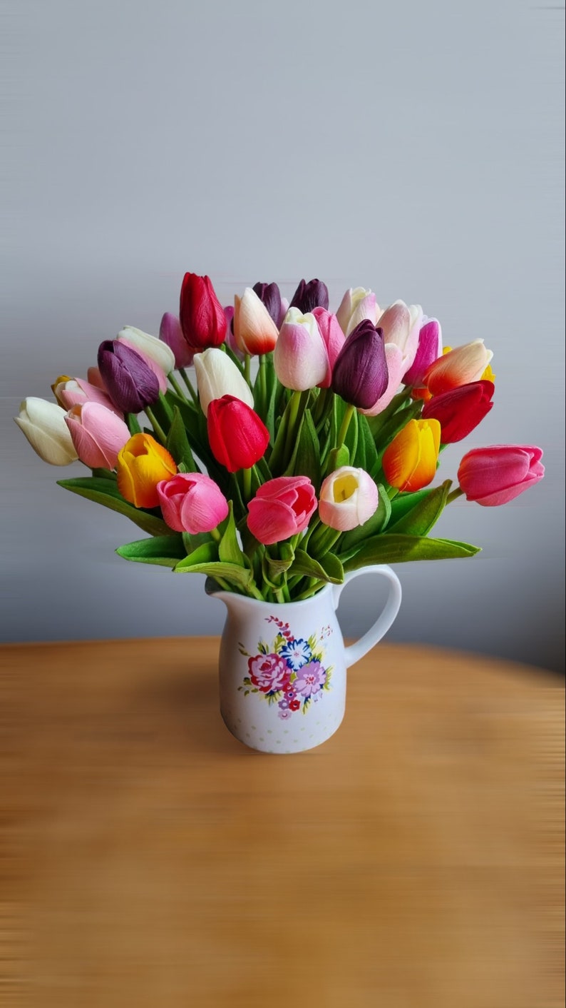 May include: A bouquet of colorful tulips in various shades of pink, purple, red, yellow, and white, arranged in a white ceramic pitcher with a floral pattern.