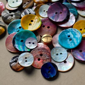 Puede incluir: Una colección de botones iridiscentes en varios tamaños y colores, incluyendo azul, morado, amarillo y rosa. Los botones tienen dos o cuatro agujeros y un acabado brillante y nacarado. Algunos botones tienen un aspecto veteado.