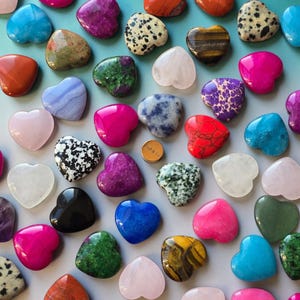 May include: A collection of heart-shaped gemstones in various colors and patterns. The gemstones include shades of pink, blue, green, and brown, with unique textures and veining. A small, gold-colored circular object is also present.