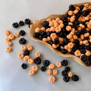 Puede incluir: Un cuenco de madera lleno de una gran colección de pequeños dulces con forma de calabaza en colores naranja y negro sólidos. Los dulces están esparcidos tanto dentro del cuenco como sobre la superficie, con crestas detalladas y forma redondeada.