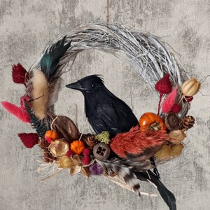 Peut inclure: Un corbeau noir est assis sur une couronne faite de brindilles et de fleurs s&eacute;ch&eacute;es. La couronne est d&eacute;cor&eacute;e d'accents rouges, orange et bruns. La couronne est suspendue &agrave; un mur gris. Le texte "Fluff & Co" est visible en bas de l'image.