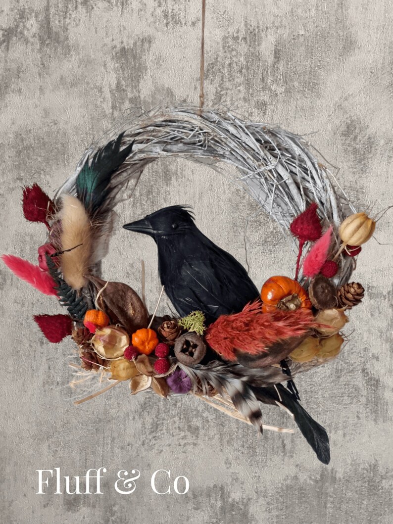 Peut inclure: Un corbeau noir est assis sur une couronne faite de brindilles et de fleurs s&eacute;ch&eacute;es. La couronne est d&eacute;cor&eacute;e d'accents rouges, orange et bruns. La couronne est suspendue &agrave; un mur gris. Le texte "Fluff & Co" est visible en bas de l'image.