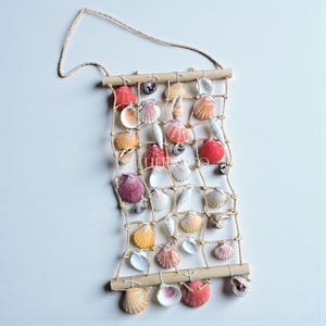 May include: A decorative wall hanging made from a rope grid, decorated with a variety of colourful seashells. The shells are in shades of red, orange, pink, and white, attached to a wooden dowel at the top and bottom, with a rope for hanging.
