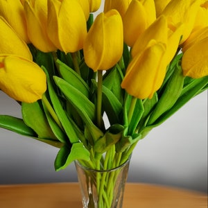 Peut inclure: Un bouquet de tulipes jaunes dans un vase en verre transparent. Les tulipes sont dispos&eacute;es en &eacute;ventail, les tiges pointant vers le centre du vase. Le vase est pos&eacute; sur une table en bois.