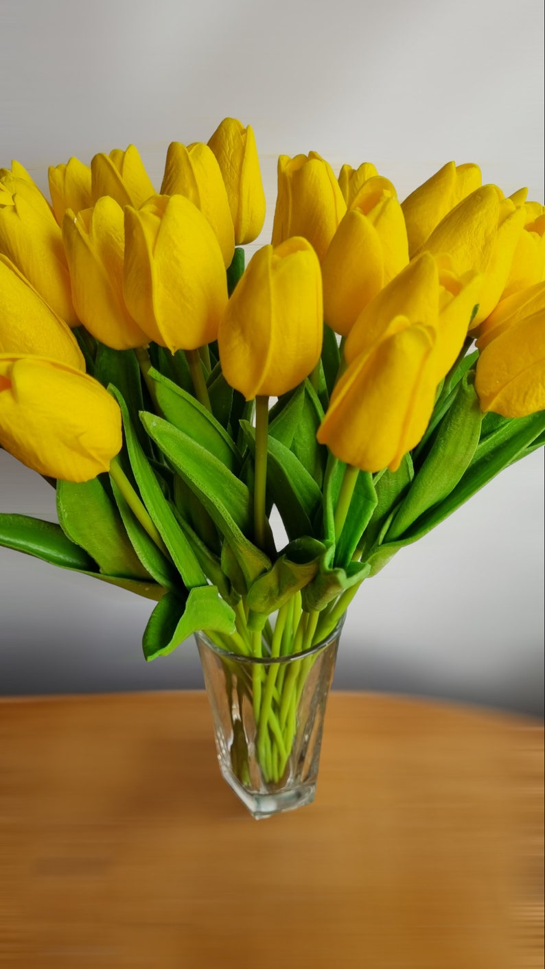 Peut inclure: Un bouquet de tulipes jaunes dans un vase en verre transparent. Les tulipes sont dispos&eacute;es en &eacute;ventail, les tiges pointant vers le centre du vase. Le vase est pos&eacute; sur une table en bois.