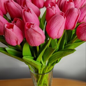 Puede incluir: Un ramo de tulipanes rosas en un jarr&oacute;n de vidrio transparente. Los tulipanes est&aacute;n dispuestos en un racimo con sus tallos extendi&eacute;ndose fuera del jarr&oacute;n.