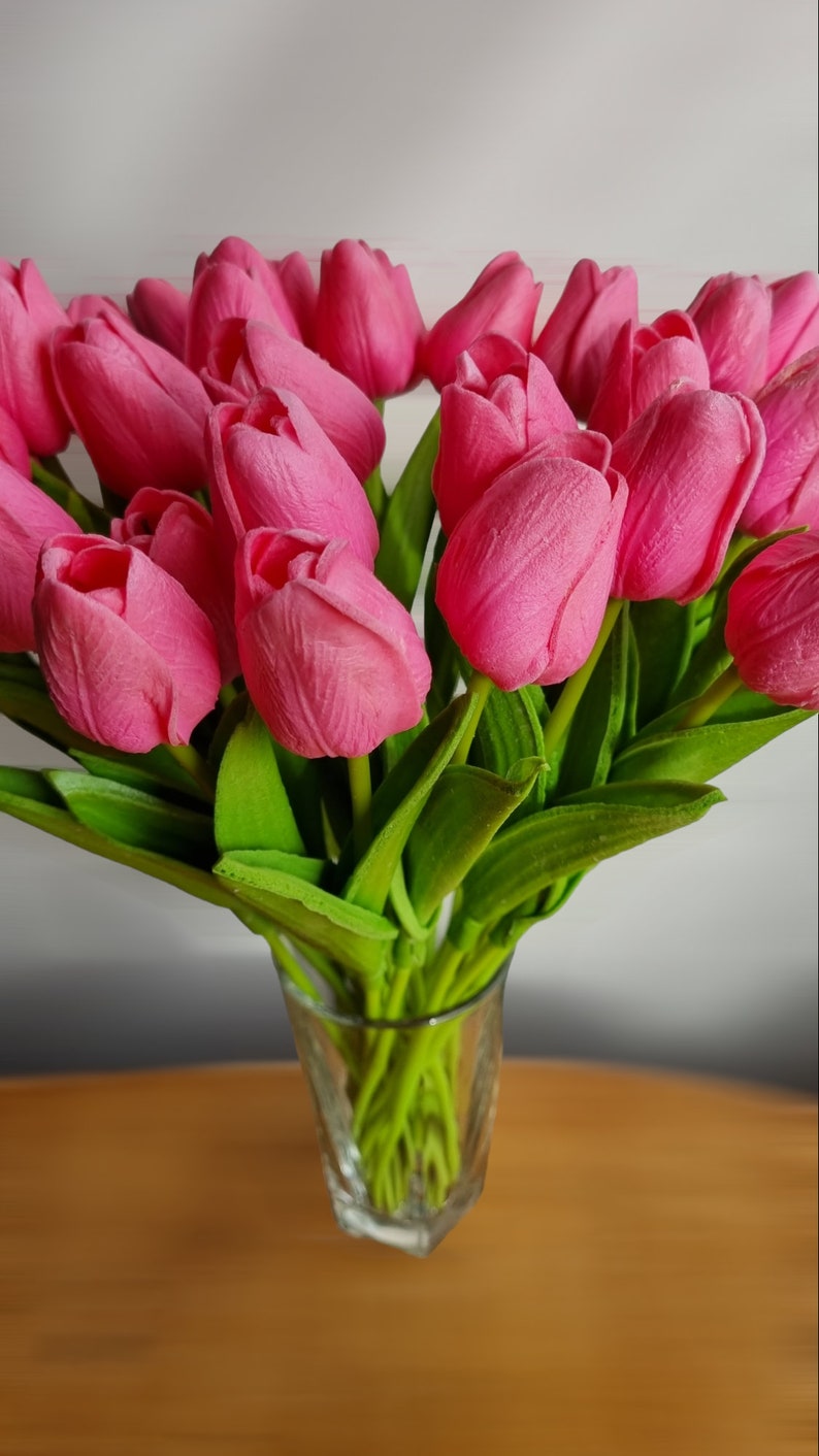 Puede incluir: Un ramo de tulipanes rosas en un jarr&oacute;n de vidrio transparente. Los tulipanes est&aacute;n dispuestos en un racimo con sus tallos extendi&eacute;ndose fuera del jarr&oacute;n.