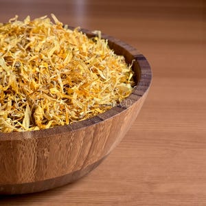 May include: A wooden bowl filled with dried, yellow calendula petals. The bowl is made of light brown wood and sits on a wooden surface. The petals are a mix of yellow and orange hues, creating a textured appearance.